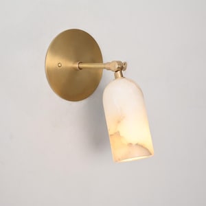 Può includere: Applique da parete con piastra posteriore circolare in ottone e braccio orientabile. La lampada ha un paralume cilindrico in materiale traslucido bianco sporco con un bagliore caldo. L'apparecchio è progettato per uso interno.