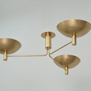 May include: A modern brass ceiling light fixture with three bowl-shaped shades. The fixture has a central stem and three arms extending to hold the shades. The light is a warm gold color and is set against a plain background.
