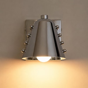 Op de afbeelding: Een gepolijste chromen wandlamp met een klokvormige kap en kleine, ronde accenten. De lamp straalt een warm licht uit. De armatuur is gemonteerd op een vierkante achterplaat. Ongeveer 20 cm hoog.