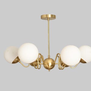 May include: A mid-century modern chandelier with a brass finish. It features five white globe shades and curved arms. The fixture is suspended from a brass rod and ceiling mount. A stylish lighting fixture for any room.