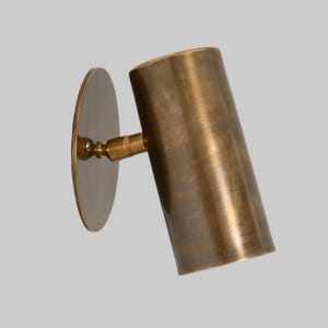 Puede incluir: Un aplique de pared cilíndrico con acabado en latón cepillado. La lámpara cuenta con una placa de montaje circular conectada a una carcasa de luz tubular mediante una pequeña articulación ajustable, mostrando una textura metálica.