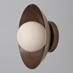 May include: A modern wall sconce featuring a spherical, off-white light bulb encased in a bronze-colored, curved metal shade. The fixture has a circular base and a minimalist design, suitable for interior lighting.
