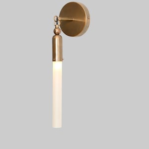 May include: A brass wall sconce with a circular backplate and a cylindrical light fixture. The light fixture is a long, slender, off-white tube. The sconce has a minimalist design, suitable for modern interiors.