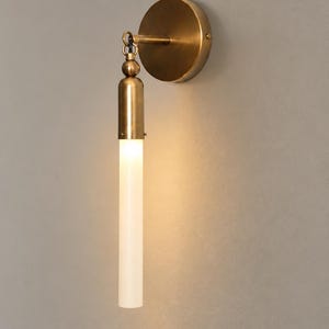 Puede incluir: Un aplique de pared con una placa circular de latón y un brazo vertical. Una pantalla cilíndrica de vidrio blanco esmerilado cuelga del brazo y está iluminada desde el interior. Los componentes metálicos tienen un acabado de latón cepillado.