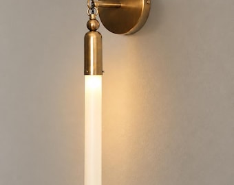 May include: A wall-mounted light fixture featuring a circular brass backplate and a vertical arm. A cylindrical, frosted white glass shade hangs from the arm, illuminated from within. The metal components have a brushed brass finish throughout.