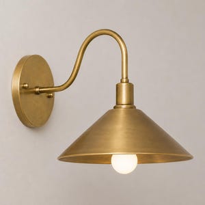 May include: A wall-mounted light fixture with a brushed brass finish. It features a circular wall plate, a curved arm extending outward, and a wide, conical shade pointing downward. A single round, frosted white light bulb is visible inside.