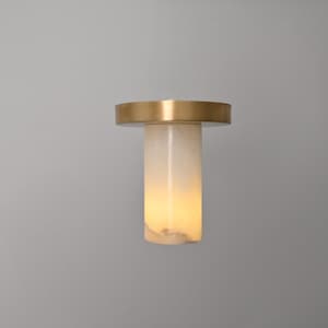 May include: A contemporary lamp featuring a brass top and a cylindrical, off-white body. The lamp's translucent form emits a warm light. The design is minimalist and elegant, suitable for modern interiors.