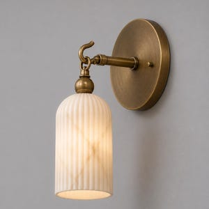 Puede incluir: Un aplique de pared con una placa circular de latón antiguo y un brazo horizontal que se conecta a un gancho. Una pantalla cilíndrica de vidrio color blanco hueso, con nervaduras verticales, cuelga del gancho mediante un remate esférico.