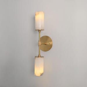 May include: A wall-mounted light fitting featuring a circular brass backplate and a vertical brass rod supporting two cylindrical, translucent white stone shades. The shades emit a soft, warm glow, and the metal components have a brushed brass finish.
