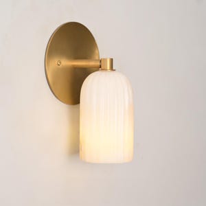 May include: A wall sconce featuring a circular brushed brass backplate and a cylindrical, vertically ribbed white glass shade. The light fitting is mounted to a wall, with the brass arm extending from the centre of the circular plate.