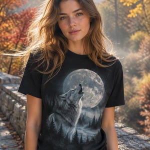 May include: A black short-sleeved t-shirt featuring a graphic print of a grey wolf howling at a large, detailed full moon. The design includes a dark forest silhouette with misty trees at the base, rendered in shades of grey, white, and black.