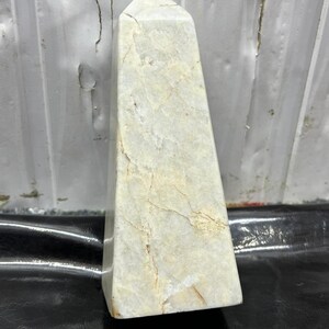 May include: A light-colored, rectangular obelisk with a pointed top. The stone has a marbled appearance with beige and cream tones, and visible natural veining. The obelisk is standing upright on a dark surface.