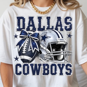 May include: A white t-shirt featuring a graphic print with the text DALLAS COWBOYS in dark blue glitter. The design includes a dark blue glitter football with a matching bow and a silver football helmet with a blue star logo and small stars.