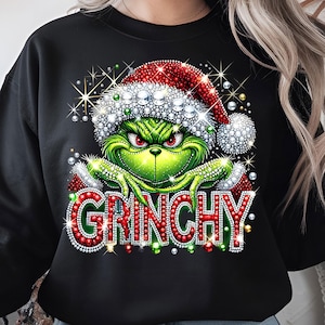 Op de afbeelding: Zwart sweatshirt met een groene Grinch-personage met een rood-witte kerstmuts. Het woord "GRINCHY" wordt weergegeven in rode en witte letters. Het ontwerp bevat sprankelende accenten.