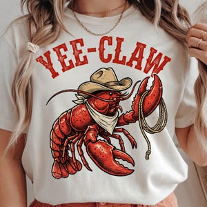 Peut inclure: T-shirt crème avec un homard rouge portant un chapeau de cowboy et un bandana, tenant une corde. Le texte "YEE-CLAW" est imprimé au-dessus du homard en rouge, dans une police de style western. Un t-shirt graphique amusant et original.