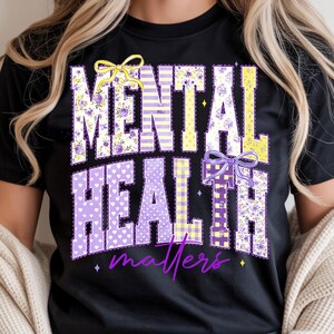 May include: Black t-shirt with the words "MENTAL HEALTH matters" in a decorative font. The letters are filled with floral, polka dot, and striped patterns in purple and yellow, with bow accents.