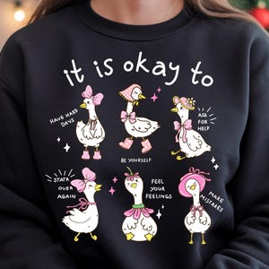 May include: Black sweatshirt with a whimsical design featuring cartoon geese with pink accessories. The text "it is okay to" is above the geese, with phrases like "Have hard days" and "Make mistakes" below.
