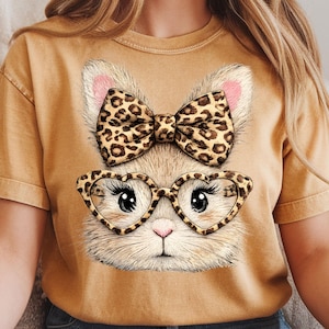 May include: Mustard-colored t-shirt featuring a cartoon bunny face with a leopard print bow and heart-shaped glasses. The bunny has pink inner ears and a small pink nose. The glasses have a leopard print pattern.
