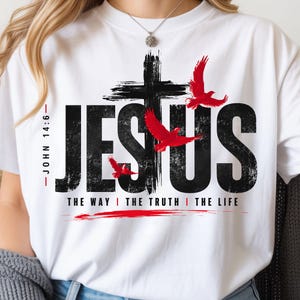 May include: A white t-shirt featuring a black distressed graphic that reads JESUS with a cross replacing the letter S. Three red birds are flying around the cross. The text JOHN 14:6 and THE WAY THE TRUTH THE LIFE are printed in black.