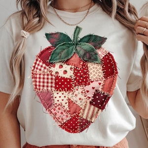 Puede incluir: Camiseta color crema con un gran diseño de fresa en patchwork. La fresa está compuesta por varias telas rojas y rosas con diferentes estampados, incluyendo cuadros, lunares y estampados florales. Hojas verdes y un tallo coronan la fresa.
