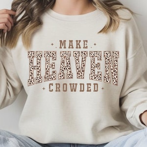 May include: A beige sweatshirt with the text "MAKE HEAVEN CROWDED" in brown letters with a leopard print pattern. The text is arched and framed by small star-like shapes.