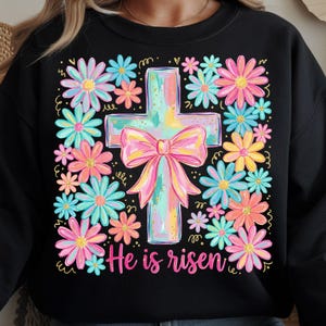 May include: Black sweatshirt with a colorful floral design surrounding a pastel cross with a bow. The words "He is risen" are written in pink script below the cross. The flowers are in shades of pink, blue, and yellow.