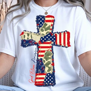 May include: White t-shirt featuring a patriotic cross design. The cross is composed of various patterns, including the American flag, camouflage, and fireworks. The words "God Bless America" are written in red script.