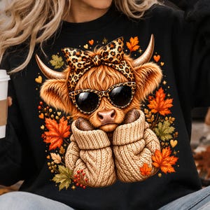 Puede incluir: Una sudadera negra con el gráfico de una vaca highland peluda que lleva un lazo con estampado de leopardo y gafas de sol. La vaca viste un suéter de punto beige, rodeada de hojas de otoño naranjas y verdes y pequeños corazones.
