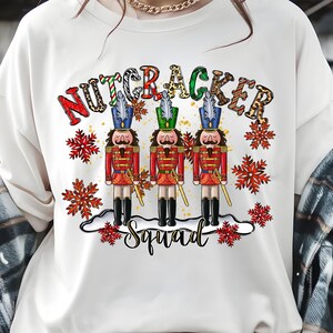 May include: Cream-colored sweatshirt featuring a festive design. The graphic includes the text "NUTCRACKER SQUAD" above three toy soldiers in red coats and black boots, with snowflake accents. The letters in "NUTCRACKER" have a variety of patterns.