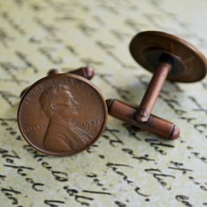 May include: A pair of cufflinks made from copper-toned US pennies. The pennies are dated 1974 and feature the profile of Abraham Lincoln. The cufflinks have a simple, classic design.