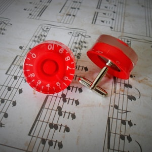 May include: Two red cufflinks, each with white numbering, designed to look like guitar amplifier knobs. They have silver-coloured posts and are displayed on a background of old sheet music.