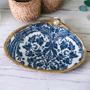 May include: A white seashell dish with a blue and gold floral pattern. The dish has a gold rim and a pearl accent.