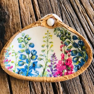 May include: A seashell dish with a floral design featuring blue, pink, and green flowers. The dish has a white background and a gold rim. A white pearl is attached to the top of the shell.