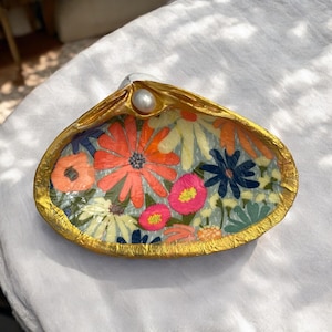 Peut inclure: Un plat en forme de coquille de mer avec un bord doré et un motif floral. La coquille est peinte avec une variété de fleurs dans des tons de rose, orange, jaune et bleu. Une perle est placée au centre de la coquille.