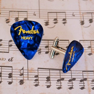 May include: A pair of cufflinks featuring a guitar pick design. The blue picks have a marbled effect and the gold text "Fender HEAVY". The cufflinks have a silver-colored metal backing and are set against a backdrop of sheet music.