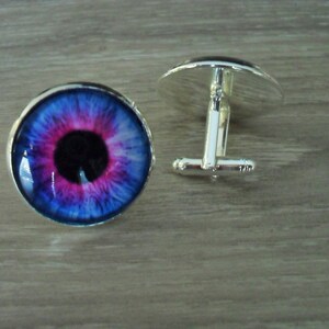 May include: A pair of silver cufflinks with a blue, pink, and black eye design.