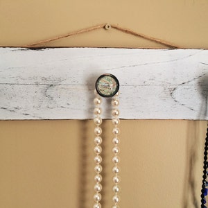 以下が含まれることがあります： 白く塗られた木製の板に、地図柄の丸いノブが付いたフックが3つ付いています。フックには、金のチェーンネックレス、真珠のネックレス、青と白のビーズのマルチストランドネックレスが掛けられています。