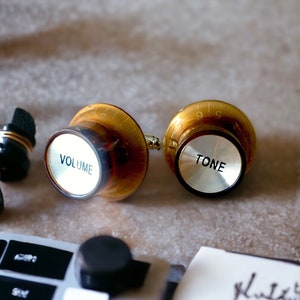 May include: Two gold and silver knobs with the words "VOLUME" and "TONE" printed on them. The knobs are on a light grey surface.