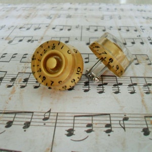 May include: A pair of cufflinks featuring circular, translucent beige knobs with black numerical markings from 0 to 10. The knobs resemble vintage audio equipment control dials, each attached to a polished silver-toned metal cufflink backing.