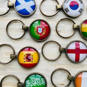 May include: A selection of keyrings displaying national flags. Each keyring has a bronze-toned metal ring and a circular emblem. Flags include Scotland, Portugal, Spain, England, Saudi Arabia, and Ireland. The flags are brightly coloured and clearly visible.