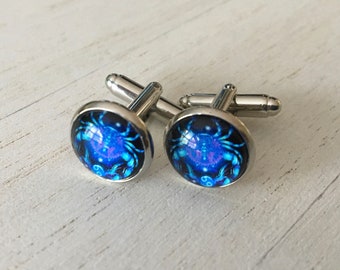 Puede incluir: Un par de gemelos de plata con un símbolo zodiacal de Cáncer azul y negro. Los gemelos son redondos y tienen un marco de metal plateado.