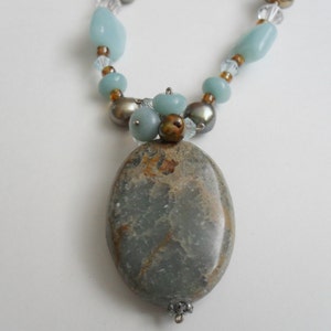 以下が含まれることがあります： 薄い青と茶色の宝石のネックレスで、大きな楕円形のペンダントが付いています。ペンダントは薄い緑と茶色の石で、大理石模様になっています。ネックレスは薄い青、茶色、透明なガラスビーズなど、さまざまなビーズで作られています。