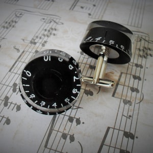 May include: A pair of cufflinks featuring black circular dials with white numbers and a silver metal connector. The dials resemble vintage amplifier knobs.