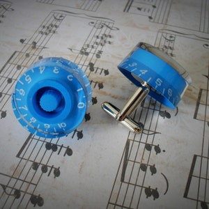 May include: A pair of cufflinks featuring blue plastic knobs with white numbers. The knobs are attached to silver metal cufflink backs.