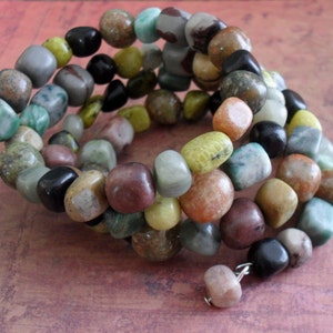 May include: Multi-colored stone bracelet with a mix of green, brown, yellow, and black stones. The bracelet is made of multiple strands of stones that are wrapped around the wrist.