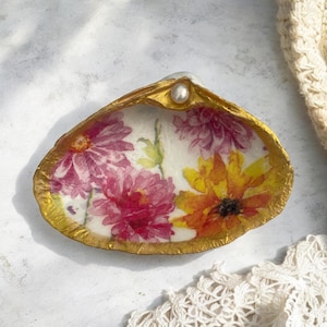 May include: A seashell dish with a gold rim and a pearl accent. The inside of the shell is decorated with a floral pattern of pink, yellow, and orange flowers.