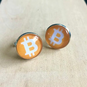 May include: A pair of silver-toned cufflinks featuring a Bitcoin symbol. Each cufflink has a round, orange background with a white Bitcoin logo. The cufflinks are displayed on a light-colored surface.