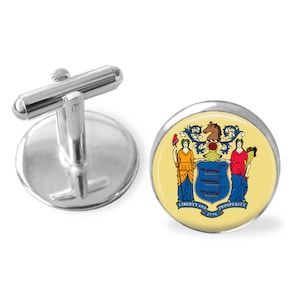 May include: Silver cufflinks with a round, yellow background featuring the state seal of New Jersey. The seal depicts a horse, a shield with three bars, and two women holding a staff and a cornucopia. The text "Liberty and Prosperity 1776" is printed below the seal.