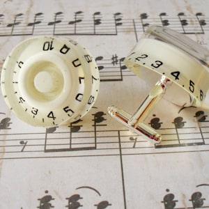 May include: A pair of cufflinks featuring white plastic guitar knobs with black numbers. The knobs are attached to silver cufflink backs.