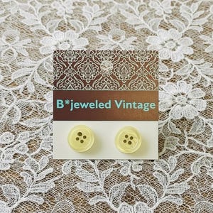 May include: Two pale yellow vintage buttons with four holes each, displayed on a white card. The card has the text "B*jeweled Vintage" and is set against a patterned background.